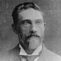 Image: Photographic head-and-shoulders portrait of a bearded, middle-aged Caucasian man. He has a full head of hair, light-coloured eyes and is wearing an Edwardian-era suit and tie