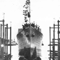 Image: A large iron warship is launched into the water. Several men stand on the bow of the ship, which is festooned in flags