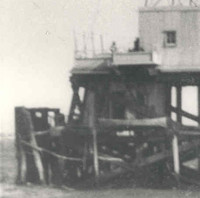 Image: A metal lighthouse with a large wooden support platform stands surrounded by water. The platform appears to be in a state of disrepair along one of its sides