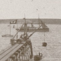 Image: A small wooden jetty projects into the ocean from a rocky coastline. A small steam vessel, four small sailboats and another island are visible in the background
