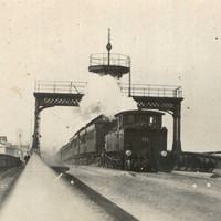 Image: A steam train passes beneath an observation tower on a bridge. A group of men in Edwardian attire perambulate along a walkway on the side of the bridge