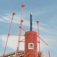 Image: The bottom section of a bright red, pre-fabricated 1860s-era lighthouse under construction. A large warehouse building is under construction in the immediate background