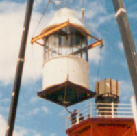 Image: A large crane hoists the lantern room of a pre-fabricated metal lighthouse to the top of the lighthouse tower. The lantern room is white and the lighthouse tower is red. A warehouse is visible in the background