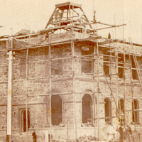 Image: An imposing, two-storey stone building under construction. The building is surrounded by scaffolding and a handful of men stand in front of its ground floor