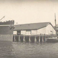 Image: A large steamship and two small sailing vessels are tied up at a wharf. A number of buildings are visible in the background