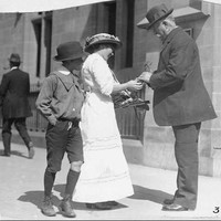 Image: A woman is selling some wattle plant to a man in the street as a young boy looks on, a man walks by in the background