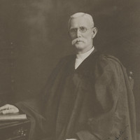 Image: Man dressed in robes, with large white moustache and glasses sits on a chair by a desk with his hand on a bible