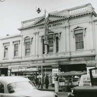 Image: Front view of a theatre building with cars driving in the foreground and people on the street