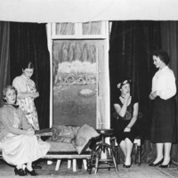 Image: Two women sitting, and two women standing on a stage with a dark curtain backdrop and various stage props