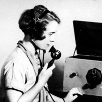 Image: A Caucasian woman in a 1930s-era dress sits at a table upon which sits a large radio set. The woman wears a headset and holds the radio receiver