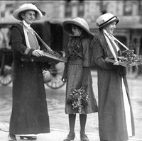 Image: Three women and a young girl wearing hats and long coats stand in the street holding sprigs of wattle plant