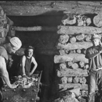 Image: Three men working inside a mine shaft with stacked timber walls and ceiling, and two trolleys filled with stone materials 