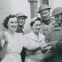 Image: Two women in white dresses hand cigarettes to a group of five men in military uniforms and helmets