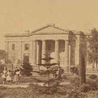 Image: three young girls walk through a garden in front of a two storey stone building with a portico featuring a triangular pediment supported by four columns