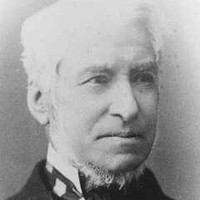 Image: a head and shoulders portrait of a man with white hair and a thin beard under his chin, his chin itself being shaven. He wears a dark coat and a striped cravat.