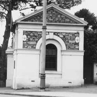Image: Small stone building with a single arched window and gable roof next to open gates. A man stands next to the gates