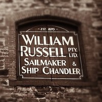 Image: Close-up photograph of part of an historic, multi-storey stone building. The words ‘Est. 1870, William Russell Pty. Ltd., Sailmaker and Ship Chandler’ are painted on the shutter of one of the building’s second-floor windows