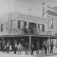 Image: group stand in front of a tavern building with four women standing on a balcony in the tavern