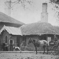 Image: man stands with dog in front of a brick house with a thatched roof. Beside him sits a woman while another man drives a horse drawn buggy.