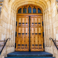 Image: The entrance to an ornate, historic building made of stone. Three large wooden doors are contained within corresponding stone archways