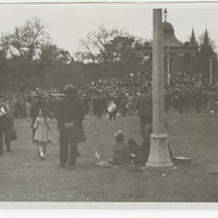 Image: a large group of men, women and children gather around a rotunda in a park