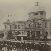 Image: A large group of people in 1880s dress stand in distinct lines in front of a large building with a domed roof which is decorated with flags and bunting. 