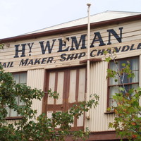 Image: The upper floor of a rectangular building with two windows and a set of double-doors. The words ‘Hy. Weman. Sail Maker, Ship Chandler’ are painted on the building front   