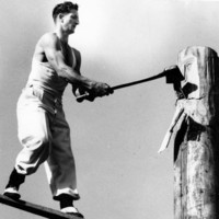 Image: man holding large axe standing on wooden plank embedded in large wooden post
