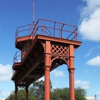 Image: A nineteenth-century iron gantry crowned by a wooden observation tower forms an archway over a modern carpark. An historic marker on a stone plinth is positioned next to the gantry
