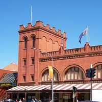 Image: A large brick building with archway windows and a square turret at one end. Cars drive by on a busy street in the foreground