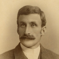 Image: A photographic head-and-shoulders portrait of a young Caucasian man wearing a late-Victorian era suit (without tie). He is sporting a large, luxurious handlebar moustache