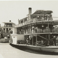 Image: Black and white photograph of two paddlesteamers on a river. Each paddlesteamer has three levels and people are visible standing and sitting on each one. The foremost paddlesteamer is flying the Union Jack flag.