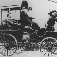 Image: A small group of Caucasian men, women and children dressed in late Victorian attire ride in a carriage-like vehicle along a town road. The vehicle has four spoked wheels and what appears to be an engine. A moustachioed middle-aged man operates it