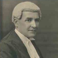 Image: A man in a barrister wig and gown poses for a photograph. He is seated in a chair with his right profile facing the camera