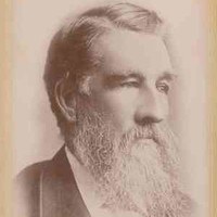 Image: A photographic head-and-shoulders portrait of a middle-aged bearded man in a suit. He has a full head of hair and is looking to the left