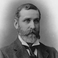Image: A photographic head-and-shoulders portrait of a bearded, middle-aged man wearing a suit and tie