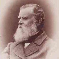 Image: A photographic head-and-shoulders portrait of a middle-aged bearded man wearing a cravat and overcoat