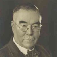 Image: A photographic portrait of a moustachioed middle-aged Caucasian man wearing wire-rimmed spectacles and a mid-20th century suit and tie