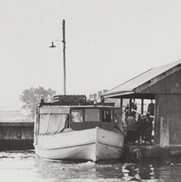 Image: A wooden motor boat with large cabin is moored alongside a jetty with a covered shelter. Several people are transiting from the jetty to the boat
