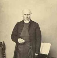 Image: Sepia photograph of bald man standing in front of a chair and table