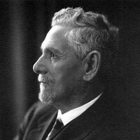 Image: A photographic head-and-shoulders portrait of a white-haired man with goatee. He is wearing a suit and is standing with his arms crossed