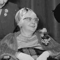 Image: A middle-aged Caucasian woman sits in a wheelchair and is wearing a fur stole and horn-rimmed glasses. Around her are seated three younger Caucasian women in formal dress