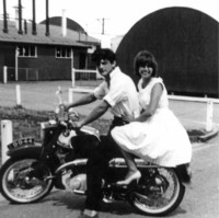 Image: Man and woman on motorbike with curved tin buildings behind