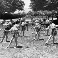 Image: A group of girls perform exercises in a grassy ground surrounded by trees and playground equipment