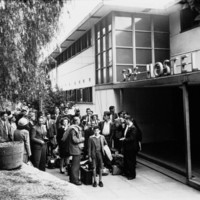 Image: A large group of British migrants of various ages arrive at hostel entrance. A sign above the door reads SPF Hostel.