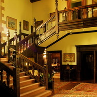 Image: An ornate wooden staircase in an historic house. Several ornate rugs and framed paintings are visible on the floors and walls surrounding the staircase
