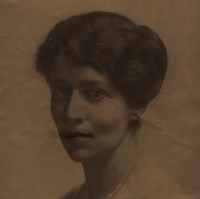 Image: A photographic head-and-shoulders portrait of a young Caucasian woman dressed in early twentieth century attire. She has dark hair and eyes and is wearing a pearl necklace and light cardigan sweater