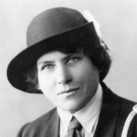 Image: A photographic head-and-shoulders portrait of a young Caucasian woman dressed in an early twentieth century Girl Guides uniform with hat. She has light-coloured eyes and dark hair