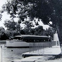 Image: a long, narrow tourist boat with a canvas roof is moored at a concrete dock with a metal railing. 
