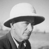 Image: A photographic head-and-shoulders portrait of a middle-aged man wearing a suit-jacket, open-necked shirt and pith helmet